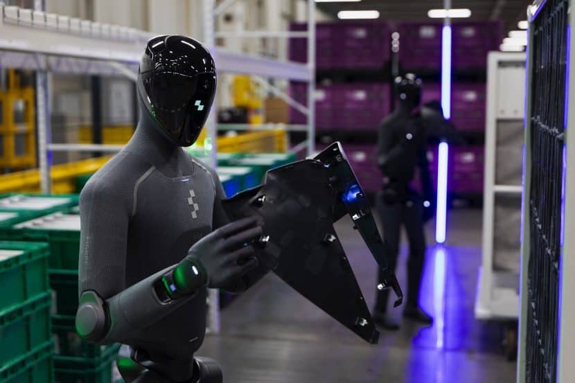 Two Figure 03 humanoid robots in a warehouse aisle. The nearer one, in a grey textile suit with a black reflective visor, holds a large, dark automotive trim panel in both hands. Green plastic bins line the shelves, and purple light rails run along the floor.
