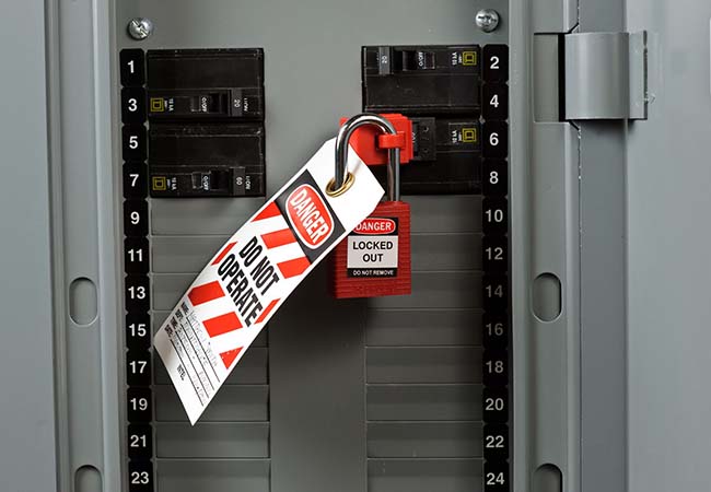 A red padlock secured through a lockout hasp on a circuit breaker in a grey electrical panel. A red and white tag reading "Danger, Do Not Operate" hangs from the hasp, and a second label reads "Danger, Locked Out."