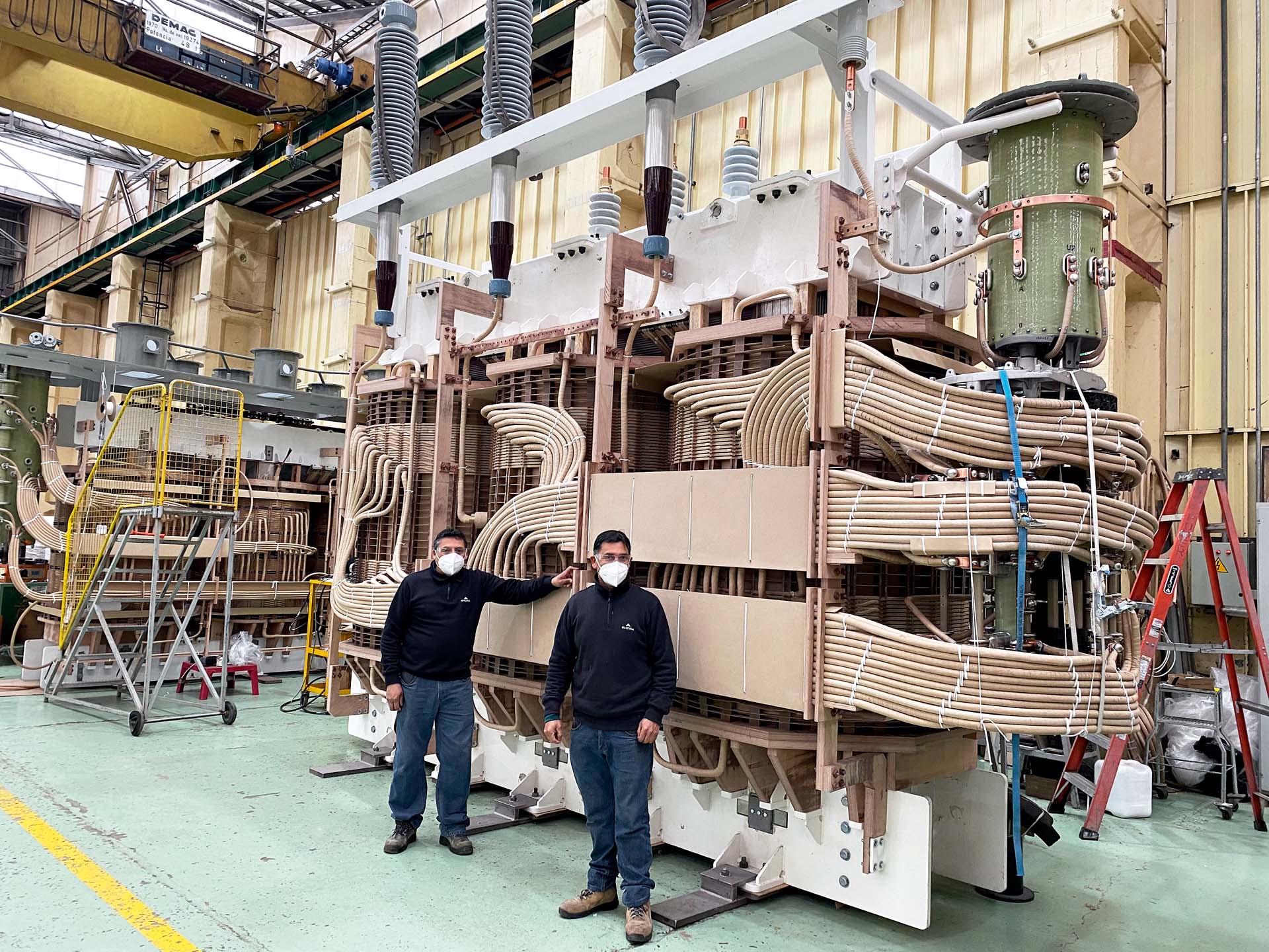 Two workers standing beside the core and coil assembly of a large power transformer on a factory floor, before it is lowered into its tank. The assembly is taller than both of them, built from stacked steel laminations with hand-formed copper conductors curving out from the windings.