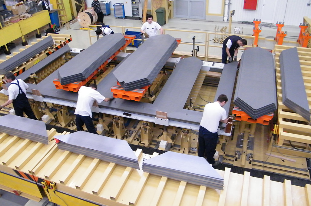 Workers on a factory floor assembling stacked sheets of electrical steel into transformer cores. Grey laminations are laid in overlapping layers along long benches, with several partially built stacks visible and staff positioning individual sheets by hand.