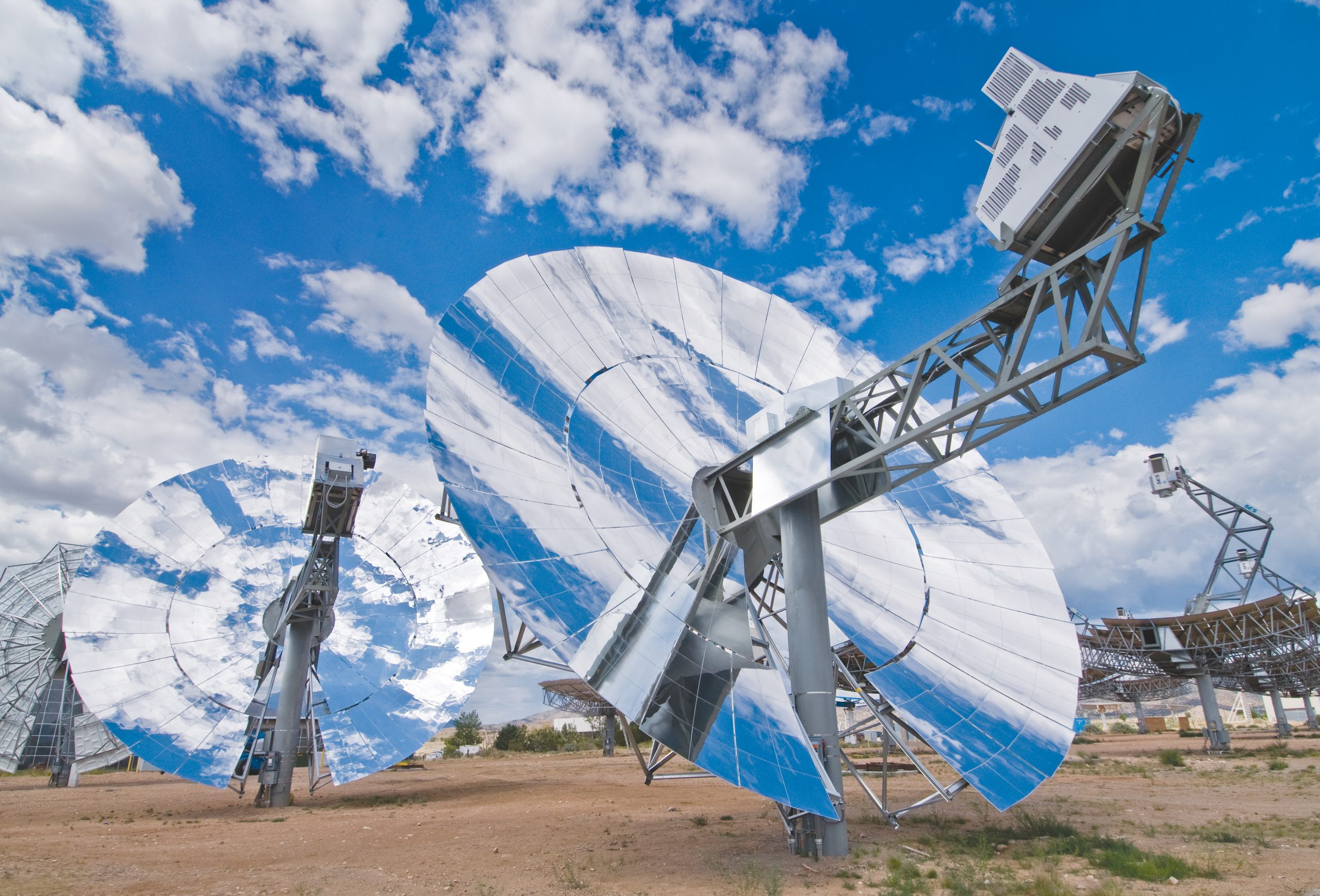 Six large parabolic dishes made of mirrored facets stand in a desert test yard under cumulus cloud, each with a power conversion unit mounted at its focal point.