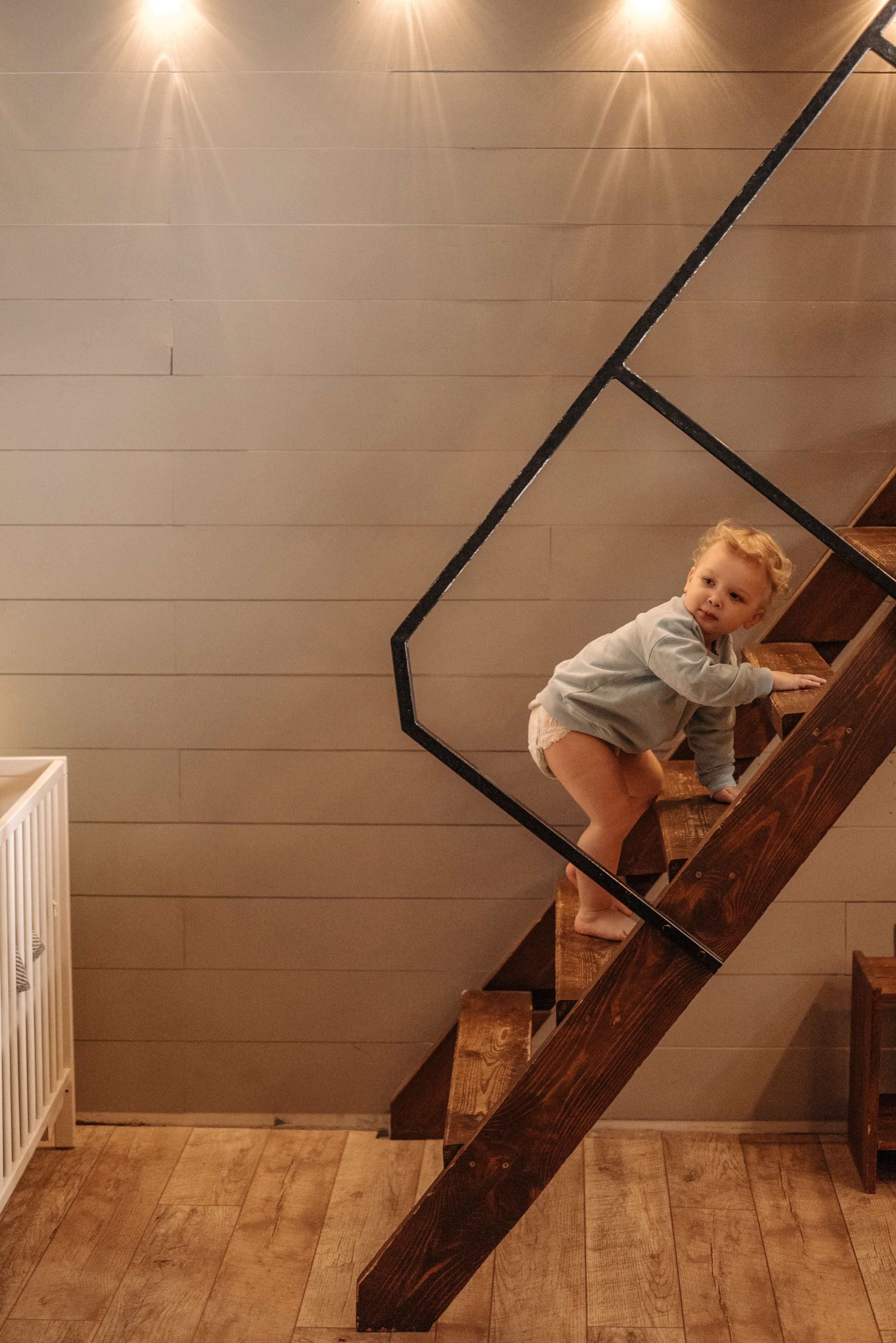 A toddler in a light blue sweater climbs an open wooden staircase with no risers, gripping a step above with one hand. A metal handrail runs well above the child's reach. A white crib stands in the corner below, on hardwood flooring.