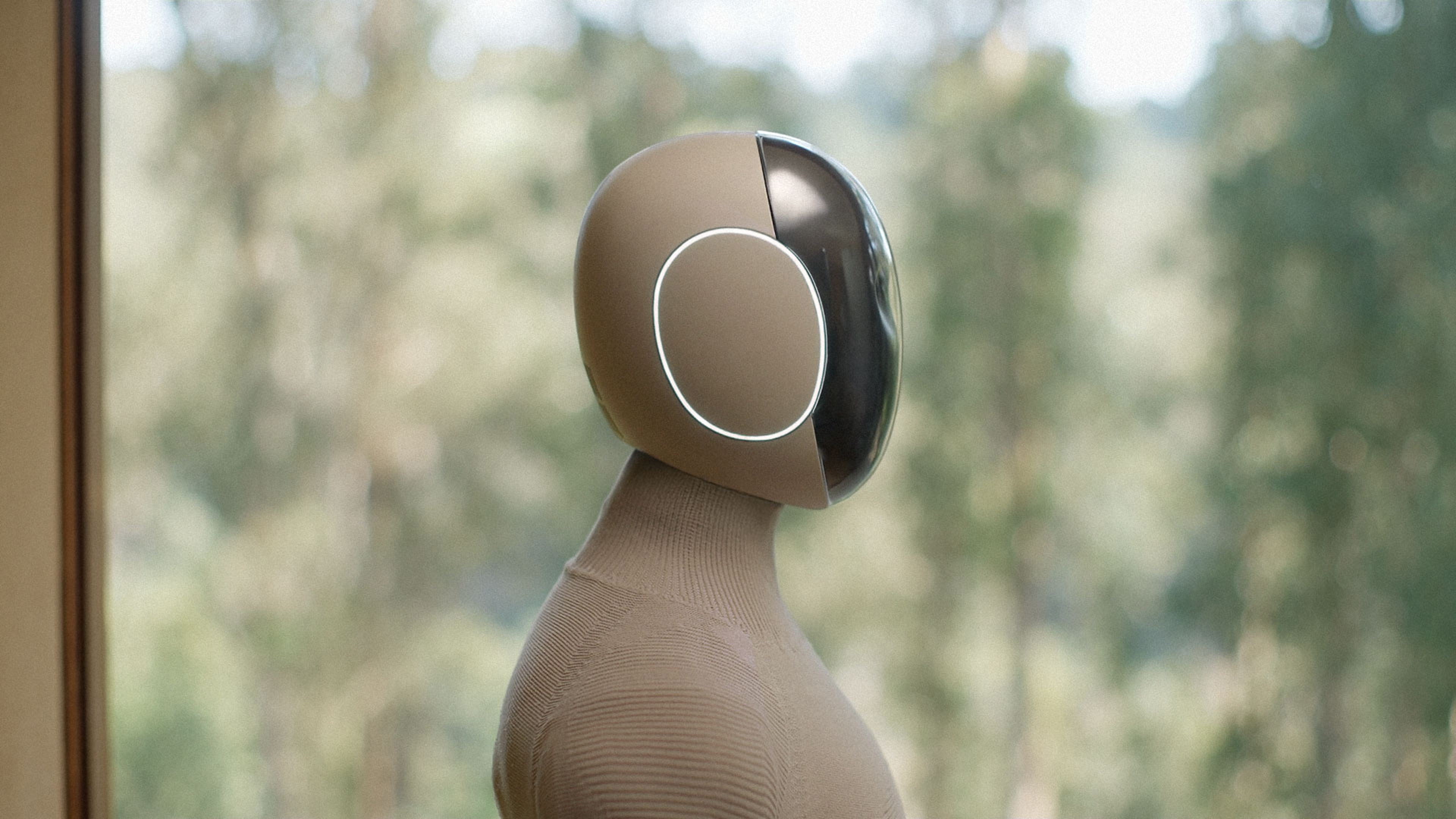 A humanoid robot with a rounded beige head and a soft knit torso, seen from the shoulders up, standing before a window with trees outside.