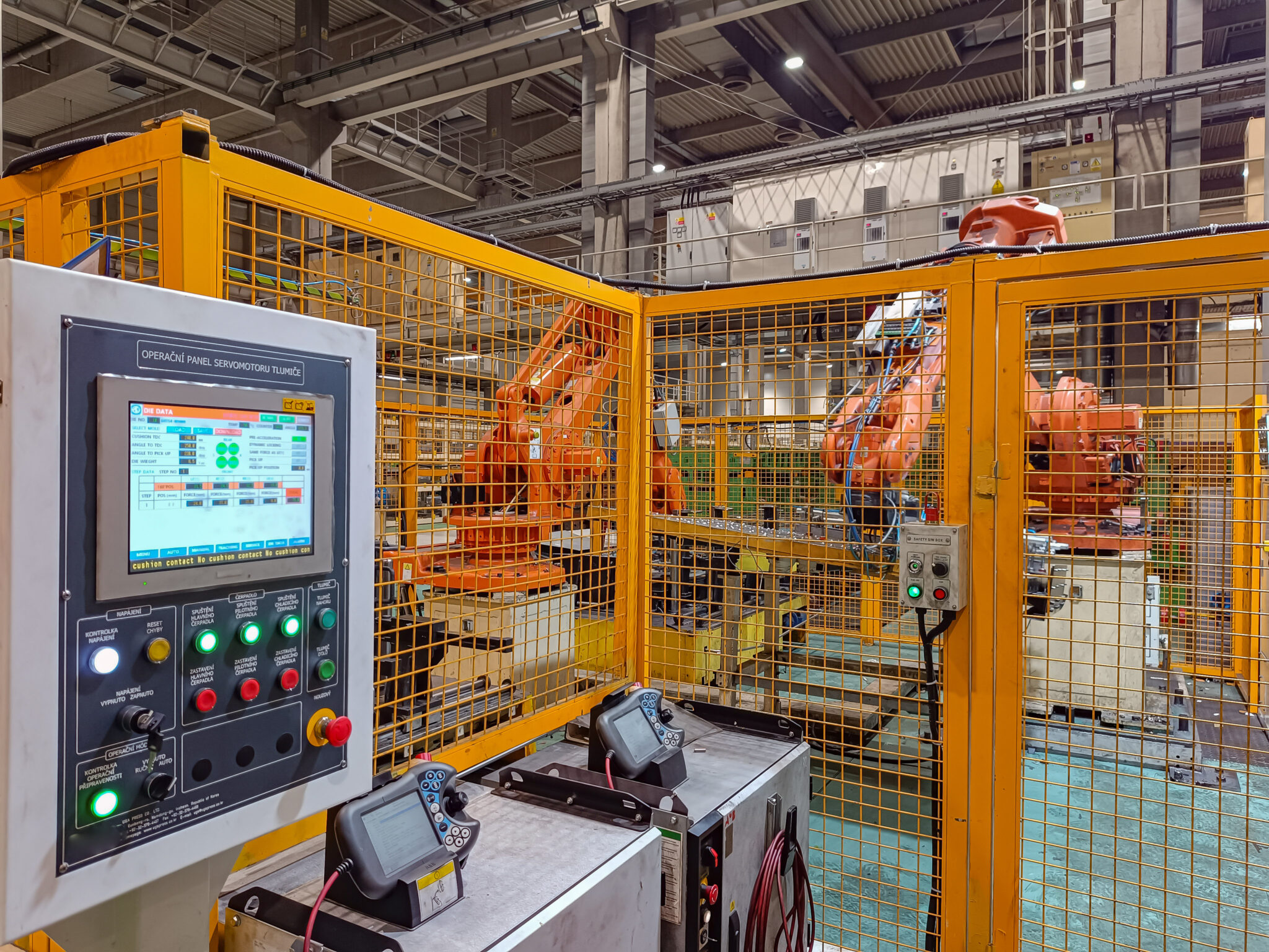 An industrial robot cell enclosed by yellow mesh safety fencing. Orange robot arms are visible inside the cage. In the foreground, a control station carries a touchscreen, indicator lights, emergency stop buttons, and two teach pendants.
