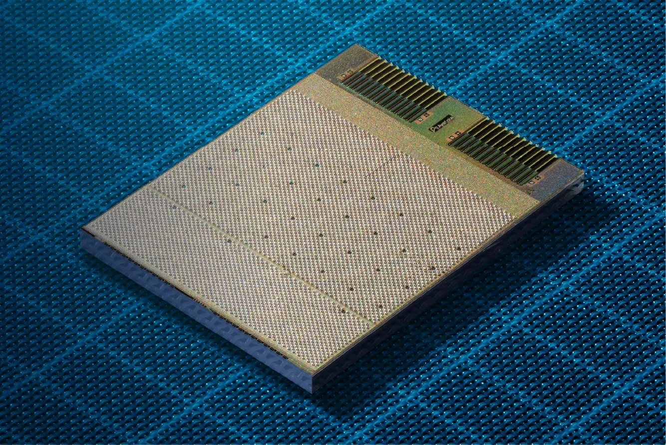 A rectangular optical I/O chiplet on a blue textured surface, showing a dense grid of contact bumps across most of its face and rows of fiber coupling structures along the top edge.