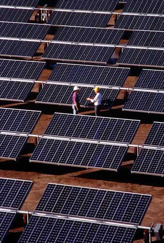 Aerial view of two workers in hard hats standing between rows of ground-mounted photovoltaic panels on bare earth.