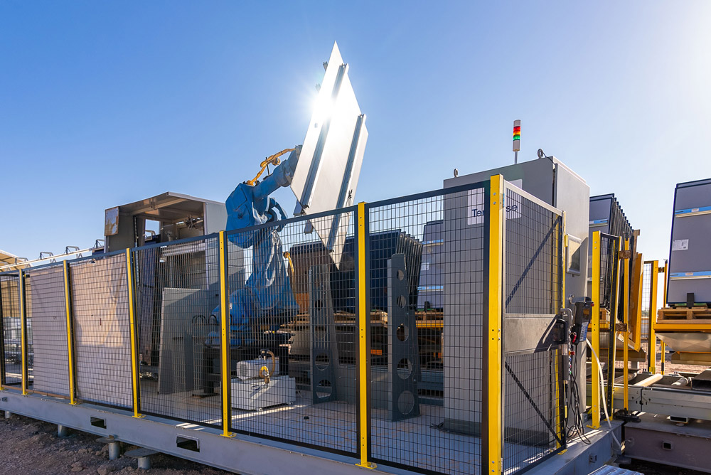 A robotic arm inside a caged assembly cell lifts a solar module against a clear sky, with yellow safety fencing, a stack light, and equipment cabinets in frame.