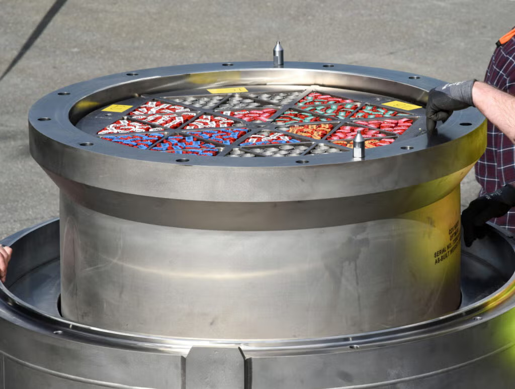 An open stainless steel transport container holding color-coded fuel compact holders, with gloved hands on its rim.